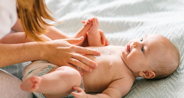 Babys wird eingecremt. Hände und Haare einer Frau sind zu sehen.