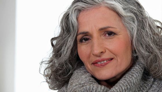 Attraktive Frau Mitte/Ende 50, schluterlange graue Haare, grauer Rollkragenpullover, schaut lächelnd in die Kamera