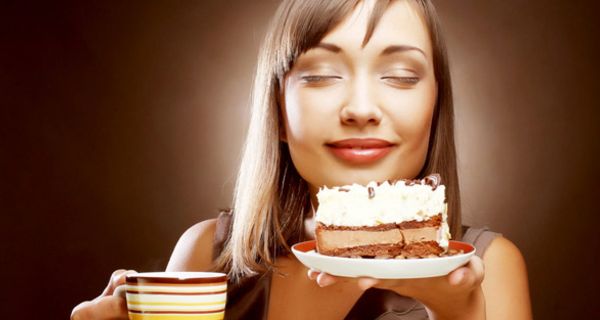Gesicht junge, braunhaarige Frau, genussvoll lächelnd, Augen geschlossen, in der linken Hand einen Teller mit einem Stück Torte, in der anderen einen Kaffeebecher