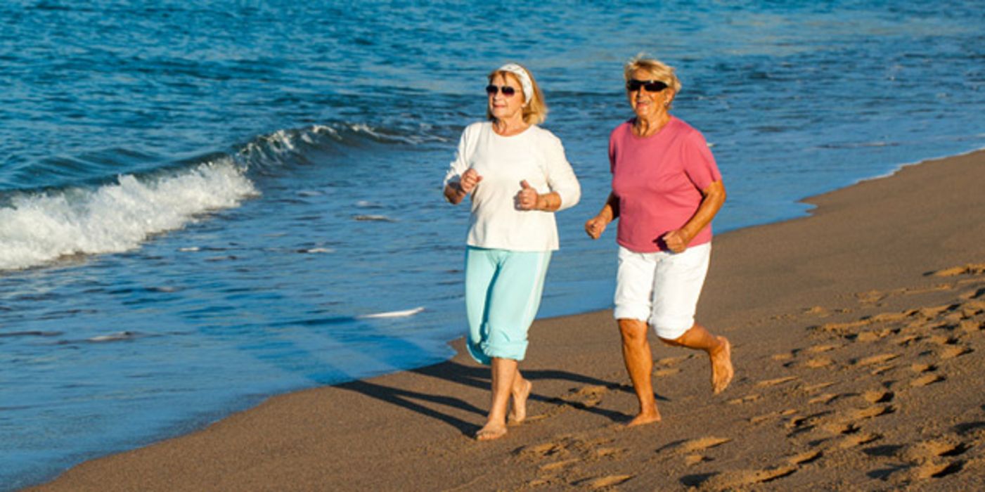 Zwei lachende Seniorinnen, sommerliche Kleidung, hochgekrempelte Hosen, Shirts, Sonnenbrillen, joggen in der Abendsonne am Strand, im Hintergrund Meereswellen