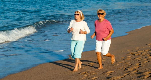 Zwei lachende Seniorinnen, sommerliche Kleidung, hochgekrempelte Hosen, Shirts, Sonnenbrillen, joggen in der Abendsonne am Strand, im Hintergrund Meereswellen