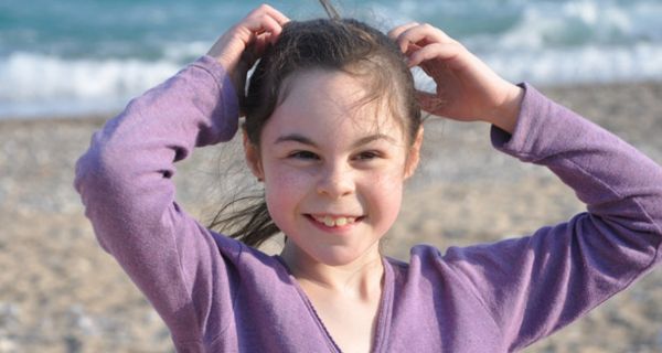 Mädchen, ca. 9 Jahre, Meer im Hintergrund, lila Feinstrickjacke, fasst sich ins Haar und blinzelt in die Kamera