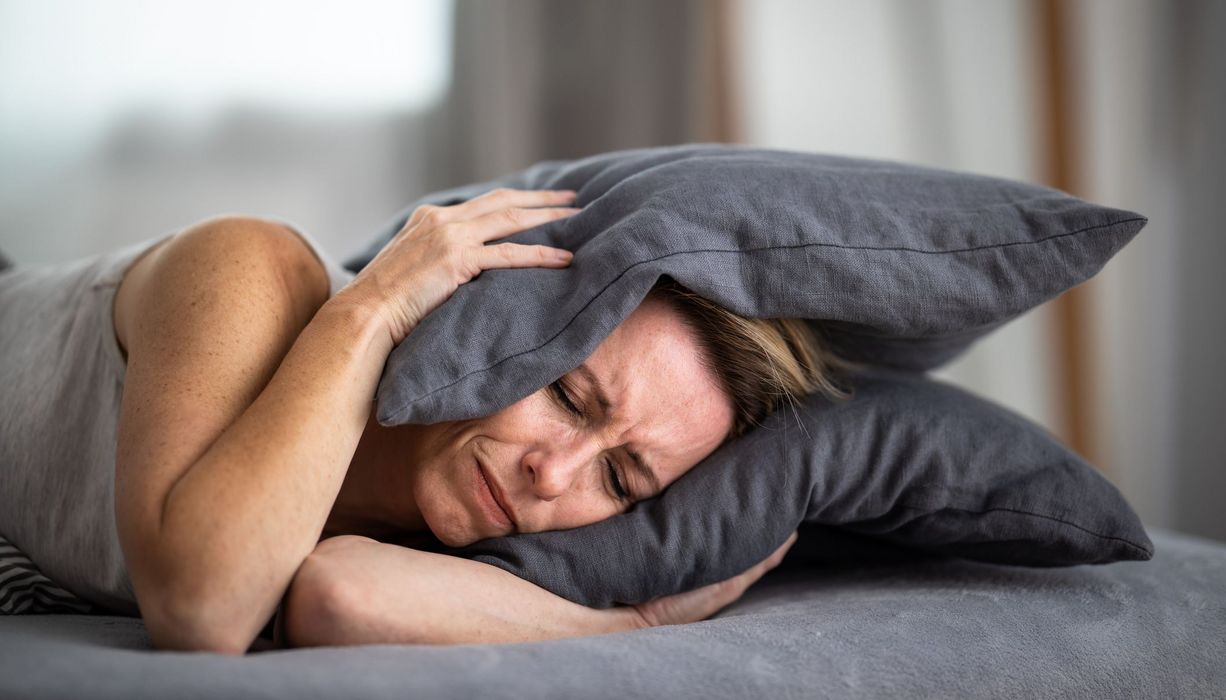 Frau, liegt im Bett und hält sich ein Kissen über die Ohren.