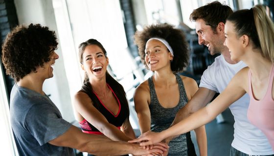 Fünf junge, heitere Menschen strecken im Fitnesstudio die Hände zusammen. Eine von ihnen lacht herzlich.