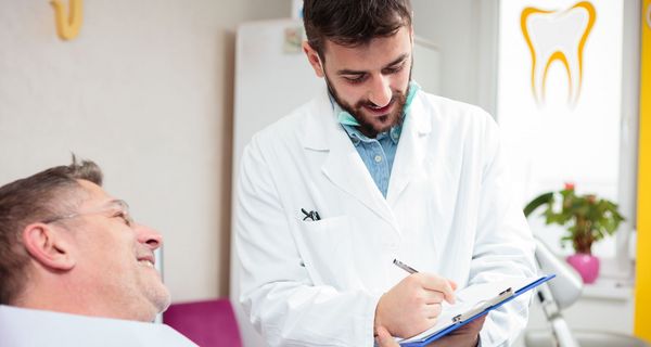 Ein jüngerer Mann im Kittel hält ein Klemmbrett in der Hand. Vor ihm sitzt ein älterer Herr der lächelt. Auf einem Fenster im Hintergrund ist ein Zahnarztpraxen-Zeichen