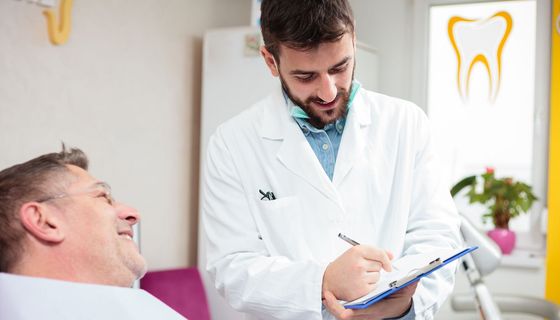 Ein jüngerer Mann im Kittel hält ein Klemmbrett in der Hand. Vor ihm sitzt ein älterer Herr der lächelt. Auf einem Fenster im Hintergrund ist ein Zahnarztpraxen-Zeichen