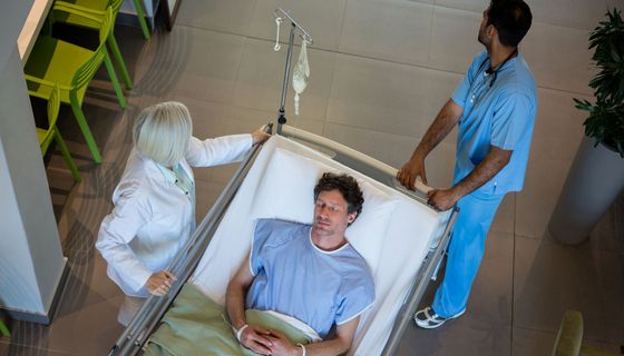 Ein Mann liegt auf einem Krankenhausbett und wird von einer Ärztin und einem Pfleger geschoben.