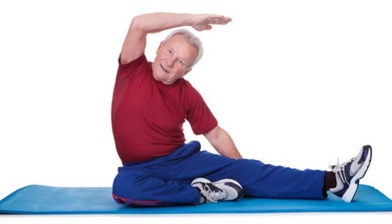 Senior sitzt mit einem seitlich ausgestreckten Bein und einem angewinkelten auf blauer Gymnastikmatte und macht eine Streckübung