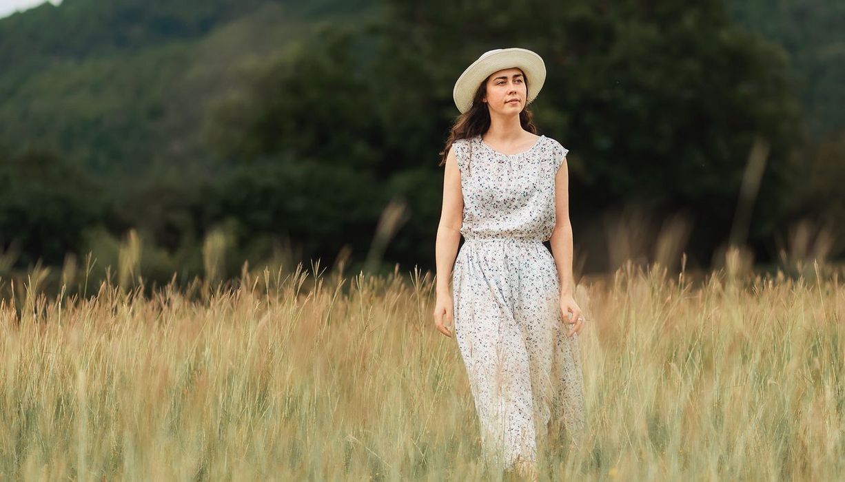 junge Frau im entspannten, weiß-gemusterten Kleid mit Sonnenhut geht im Feld spazieren. 