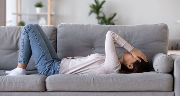 Junge Frau, liegt auf der Couch, hält ihre Hände an den Kopf.