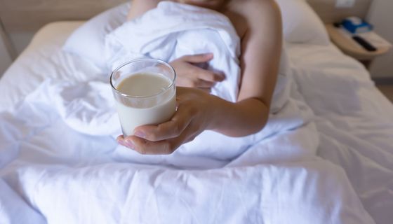 Frau, sitzt im Bett und hält ein Glas Milch in der Hand.