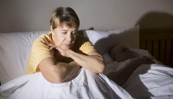 Eine ältere Frau im gelben Oberteil sitzt aufrecht neben einer schlafenden Person im Bett.