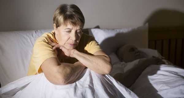 Eine ältere Frau im gelben Oberteil sitzt aufrecht neben einer schlafenden Person im Bett.