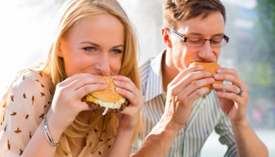 Junge Frau und junger Mann sitzen in ihrer Mittagspause im Freien und beißen herzhaft in einen Hamburger