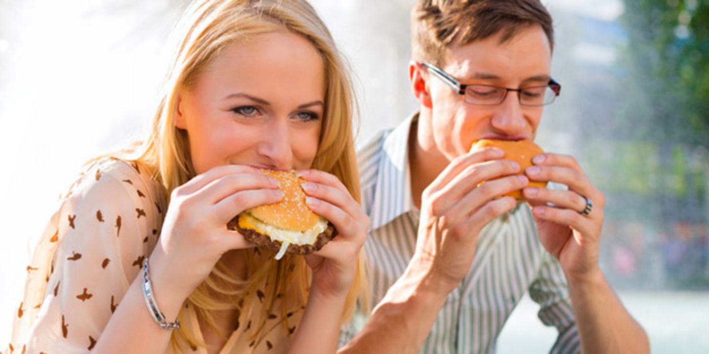Junge Frau und junger Mann sitzen in ihrer Mittagspause im Freien und beißen herzhaft in einen Hamburger