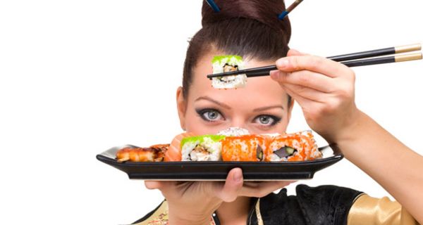 Nahaufnahme asiatisch anmutender Frau, die Sushiteller vors Gesicht hält und ein Sushi mit Stäbchen fasst.