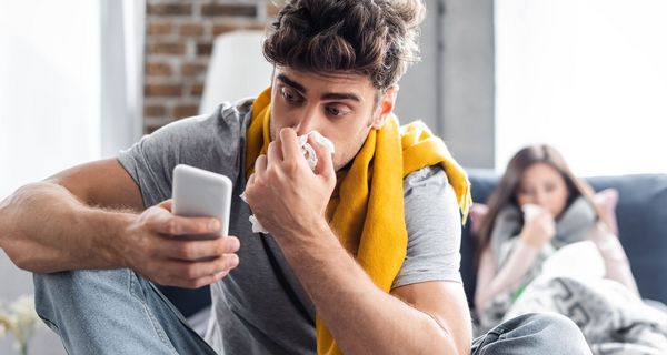 Junger Mann, putzt sich die Nase und schaut auf sein Handy.