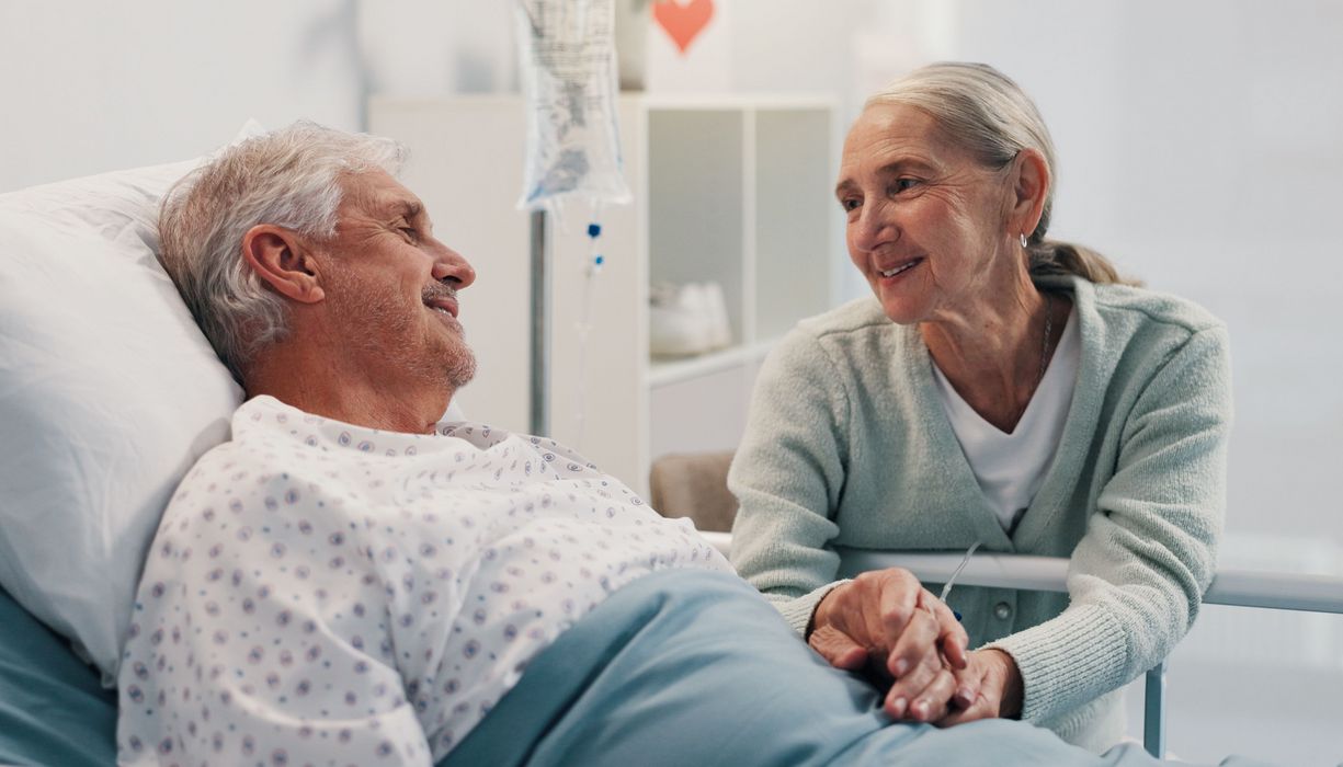 Ein älterer Mann liegt im Krankenhaus, eine Frau im gleichen Alter hält seine Hand. Beide lächeln sich an.