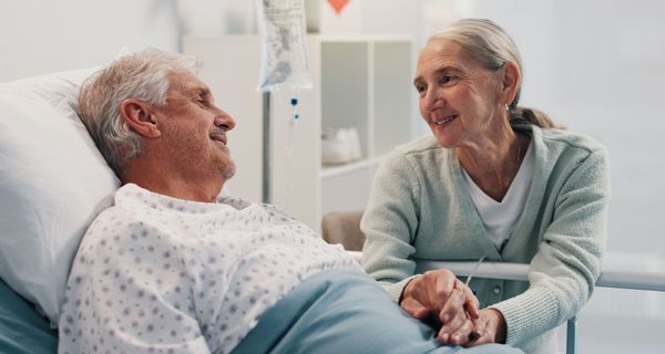 Ein älterer Mann liegt im Krankenhaus, eine Frau im gleichen Alter hält seine Hand. Beide lächeln sich an.
