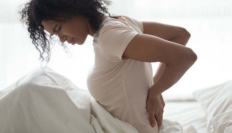 Junge Frau fasst sich im Bett an unteren Rücken, schmerzverzerrter Gesichtsausdruck