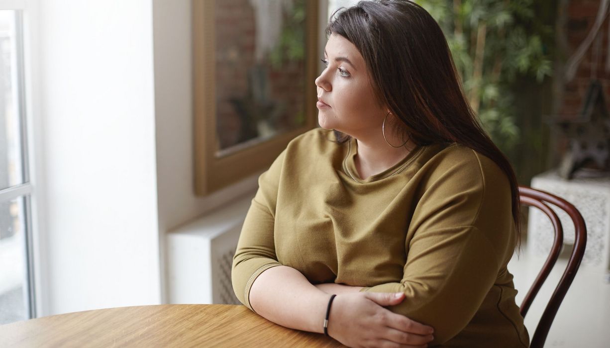 Junge übergewichtige Frau, sitzt an einem Tisch und schaut nach draußen.