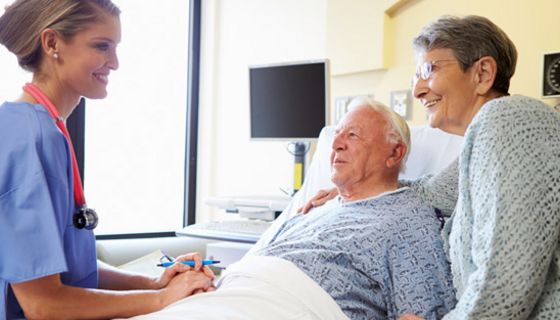 Älterer Mann im Krankenhausbett, Frau steht daneben, beide unterhalten sich lächelnd mit einer jungen Krankenschwester