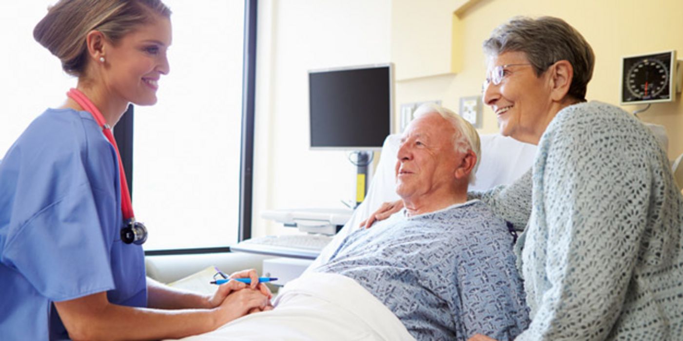 Älterer Mann im Krankenhausbett, Frau steht daneben, beide unterhalten sich lächelnd mit einer jungen Krankenschwester