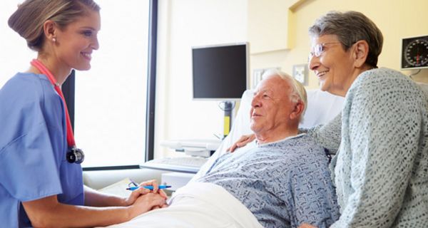 Älterer Mann im Krankenhausbett, Frau steht daneben, beide unterhalten sich lächelnd mit einer jungen Krankenschwester