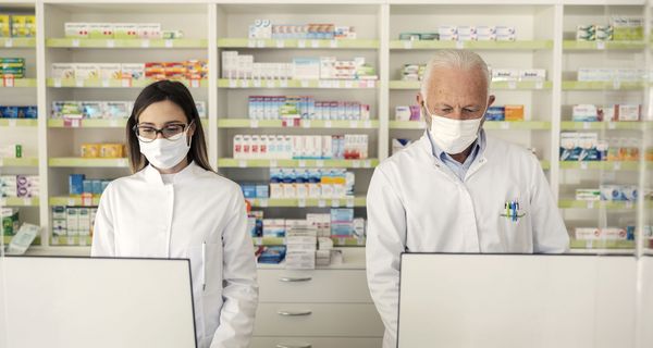 Eine Apothekerin und ein Apotheker mit Maske vor großen Bildschirmen.