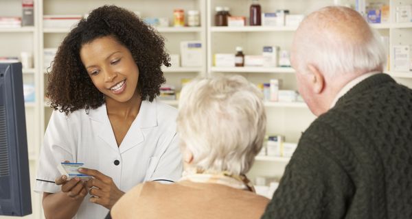 Eine junge Apothekerin hält ein Arzneimittel in der Hand und erklärt einem älteren Paar etwas.