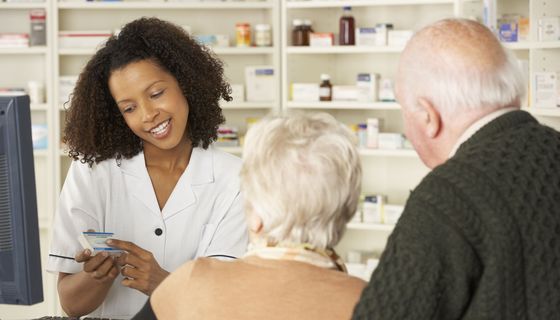 Eine junge Apothekerin hält ein Arzneimittel in der Hand und erklärt einem älteren Paar etwas.