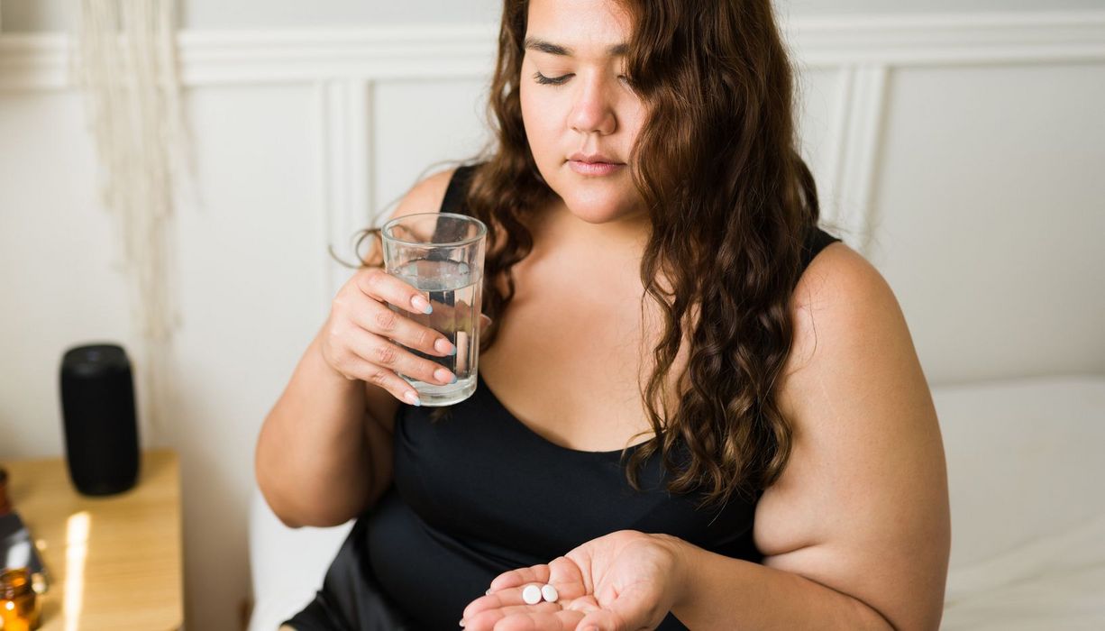 Junge Frau mit Übergewicht blickt zweifelnd auf Tabletten in ihrer Hand. In der anderen hält sie ein Wasserglas.