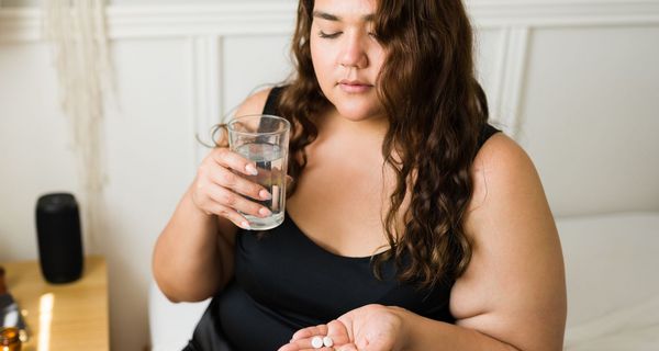 Junge Frau mit Übergewicht blickt zweifelnd auf Tabletten in ihrer Hand. In der anderen hält sie ein Wasserglas.