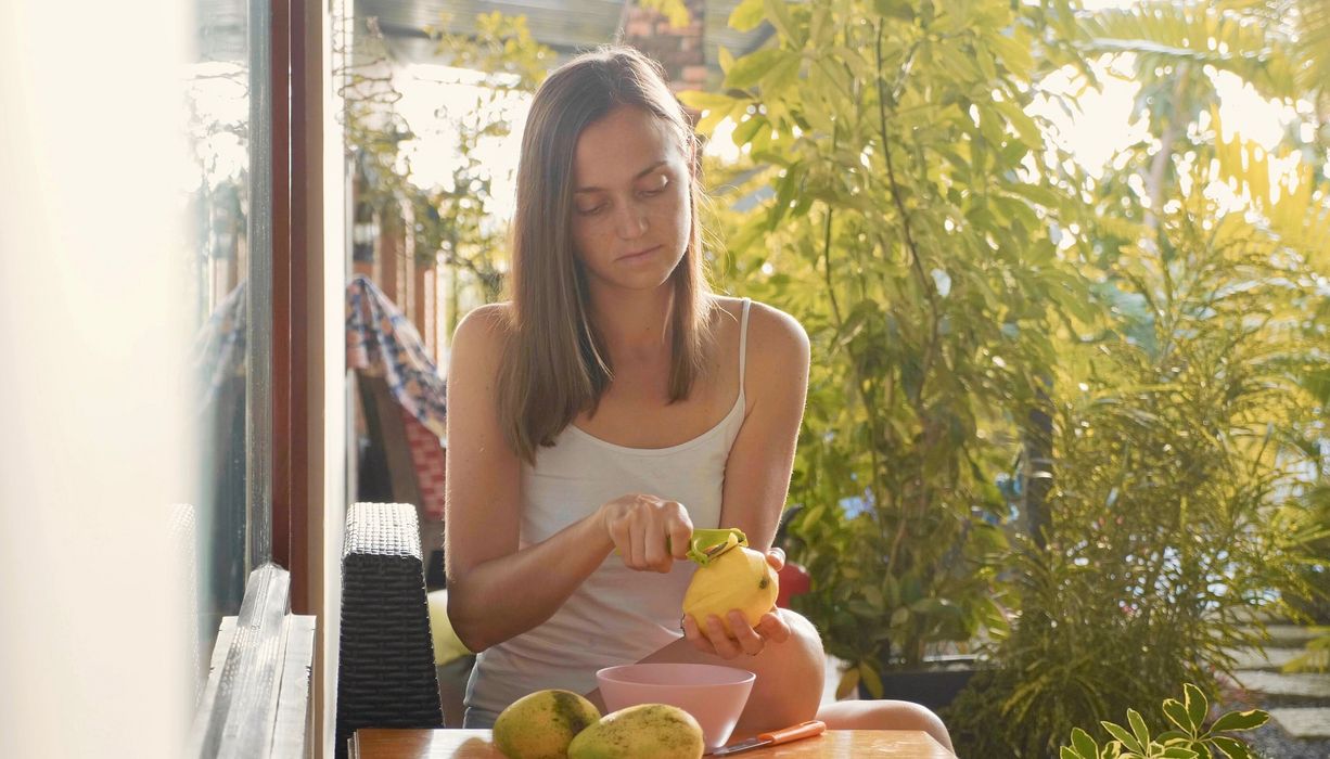 Eine Frau sitzt an einem Tisch im Garten und schält Mangos.