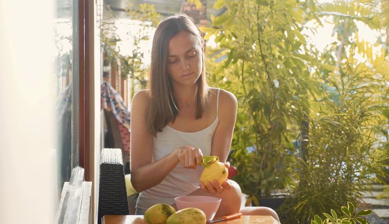 Eine Frau sitzt an einem Tisch im Garten und schält Mangos.