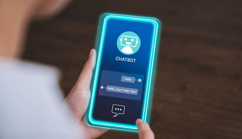 Blick auf ein Smartphone mit einer geöffneten Chatbot-App: Der Bot hat geschrieben: "Hello" und "Can I help you?"
