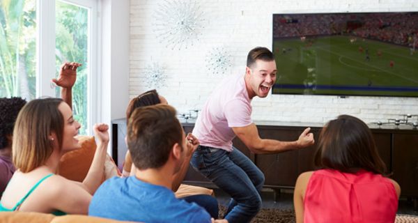 Gruppe junger Menschen schaut im Wohnzimmer ein Fußballspiel im Fernsehen, ein junger Mann steht, ballt die Faust und feuert seine Mannschaft an
