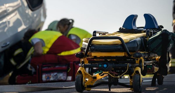 Im Vordergrund: Eine spezielle Liege mit Stütze für den Nacken, im Hintergrund: Ein Unfallwagen auf dem Dach, Sanitäter und Feuerwehr in der Hocke vor dem Fenster.