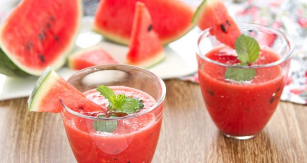 Wassermelonensaft in 2 Gläsern, dahinter aufgeschnittene Wassermelonen