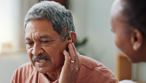 Älterer Herr fasst an sein Hörgerät, unscharf im Vordergrund: Der Kopf einer Krankenschwester von der Seite.