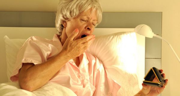 Ältere Frau im Bett gähnt und schaut auf einen Wecker.