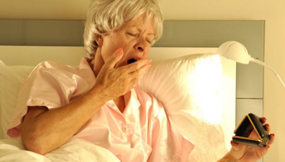 Ältere Frau im Bett gähnt und schaut auf einen Wecker.