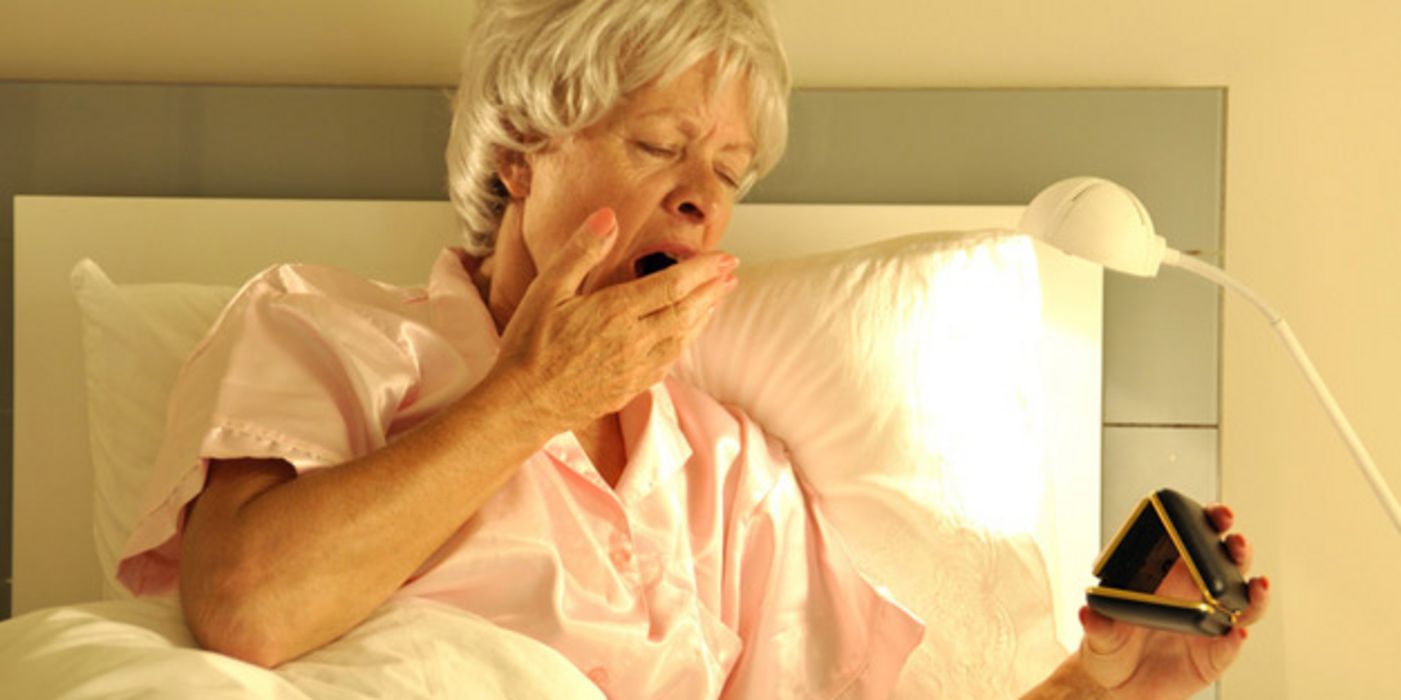 Ältere Frau im Bett gähnt und schaut auf einen Wecker.
