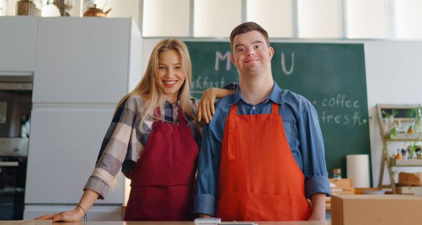 Junger Mann mit Down-Syndrom steht neben einer jungen Frau, beide tragen rote Kochschürzen.