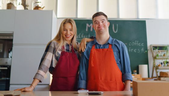 Junger Mann mit Down-Syndrom steht neben einer jungen Frau, beide tragen rote Kochschürzen.