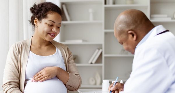 Schwangere Frau, eine Hand auf ihren Bauch gelegt, sitzt einem Arzt gegenüber, der sich Notizen macht.
