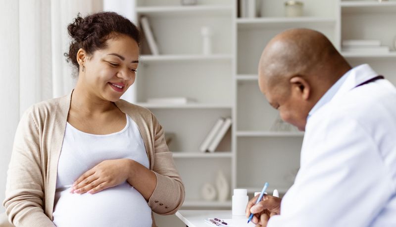 Schwangere Frau, eine Hand auf ihren Bauch gelegt, sitzt einem Arzt gegenüber, der sich Notizen macht.