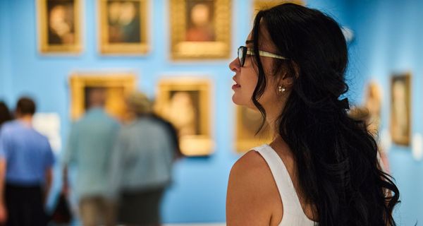 Im Vordergrund: Eine junge Frau, die nach links aus dem Bild heraus blickt, im Hintergrund (unscharf): Blaue Wände mit historischen Portraits und eine kleine Gruppe Museumsbesucher.