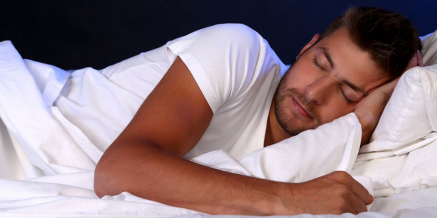 Jüngerer dunkelhaariger Mann schläft in Seitenlage in weißbezogenem Bett, nachtblauer Hintergrund