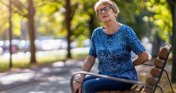 Ältere Frau, sitzt auf einer Parkbank und hält eine Hand an den unteren Rücken.